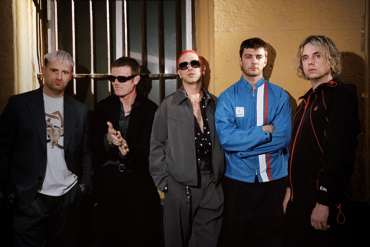 Post-punk band Fontaines D.C. standing together in dark streetwear and jackets against a yellow wall, photo by Ewen Spencer.